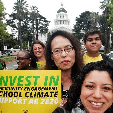 Public Advocates Of Counsel Liz Guillen holds a sign that says "Invest in Community Engagement, School Climate, Support Assembly Bill 2820". Students and family leaders are smiling in the background.