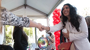 A woman smiling and shaking the hand of a person who is not in the frame of the photograph. They are under an outdoor tent with a balloon arch and a prize wheel in the background.