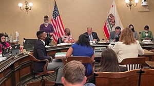 A group of people, some masked, sit inside a courtroom. There is an American flag and a Californian flag behind the judges.